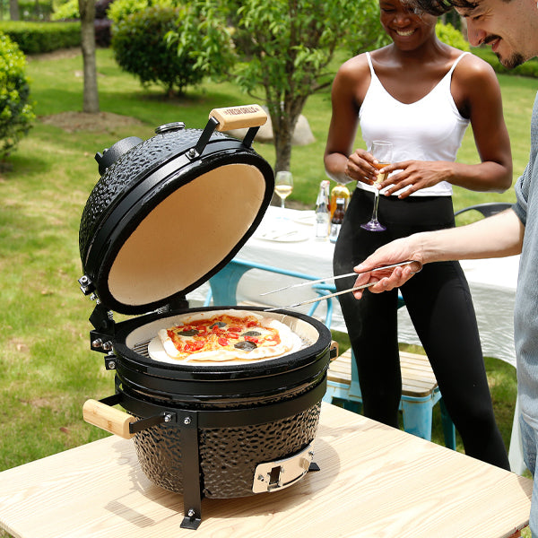 Kamado-Ofen Holzkohlegrill in Schwarz mit Holzgriffen, geöffnet und bereit zum Grillen, während eine Person Pizza zubereitet und eine Frau zuschaut