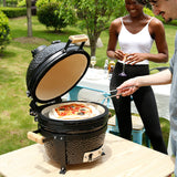 Kamado-Ofen Holzkohlegrill in Schwarz mit Holzgriffen, geöffnet und bereit zum Grillen, während eine Person Pizza zubereitet und eine Frau zuschaut