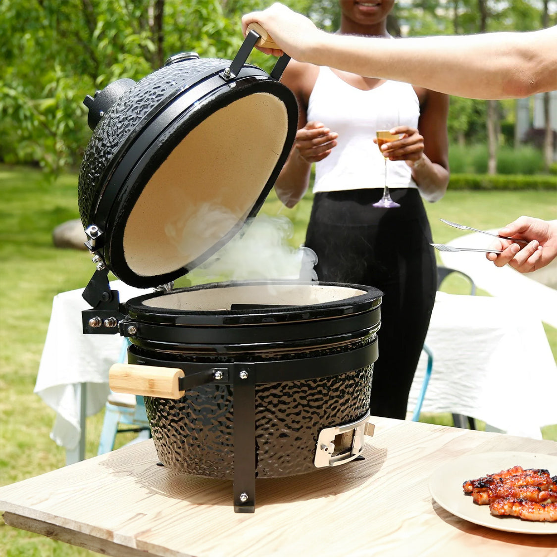 Schwarzer Kamado-Ofen mit Holzgriff, geöffnet, dampfend, auf einem Holztisch im Freien, umgeben von grünem Gras und einer Person mit Getränk