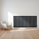 Schwarzer Flachheizkörper mit vertikalen Lamellen, 600mm x 1190mm, elegant an einer hellen Wand, neben einem modernen weißen Sessel auf Holzfußboden