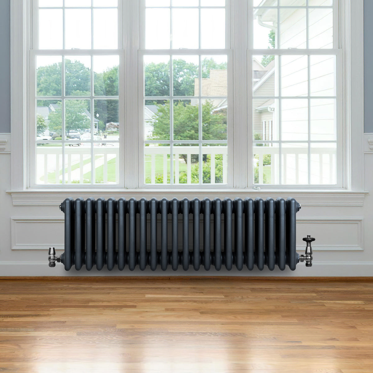 Röhrenheizkörper mit 4 Säulen in Anthrazitgrau, ausgestattet mit Chromventilen, vor einem großen Fenster auf einem Holzfußboden