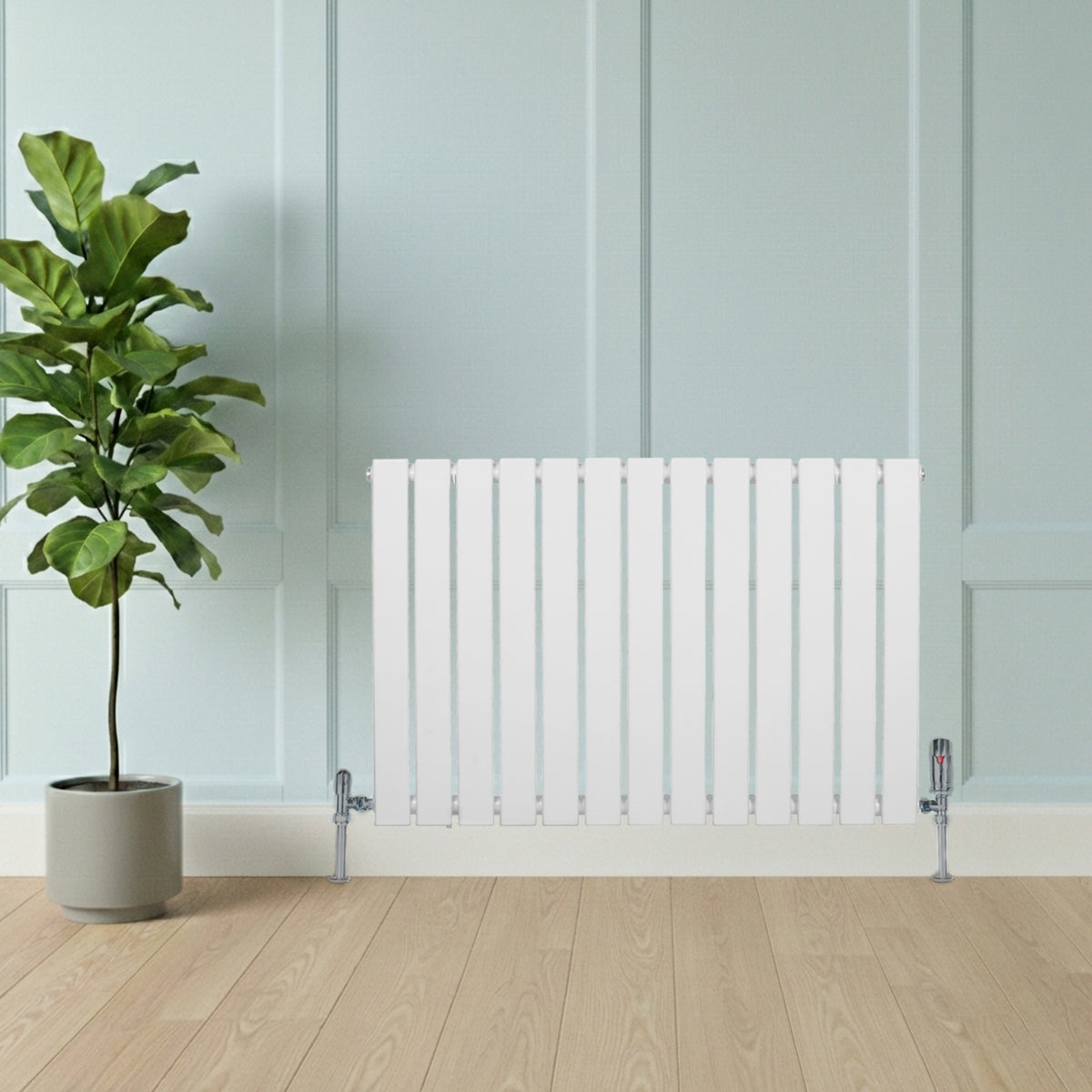 Flachheizkörper in Weiß, 600mm x 910mm, mit vertikalen Lamellen und glänzenden Ventilen, vor einer hellen Wand und einem grünen Pflanzenarrangement