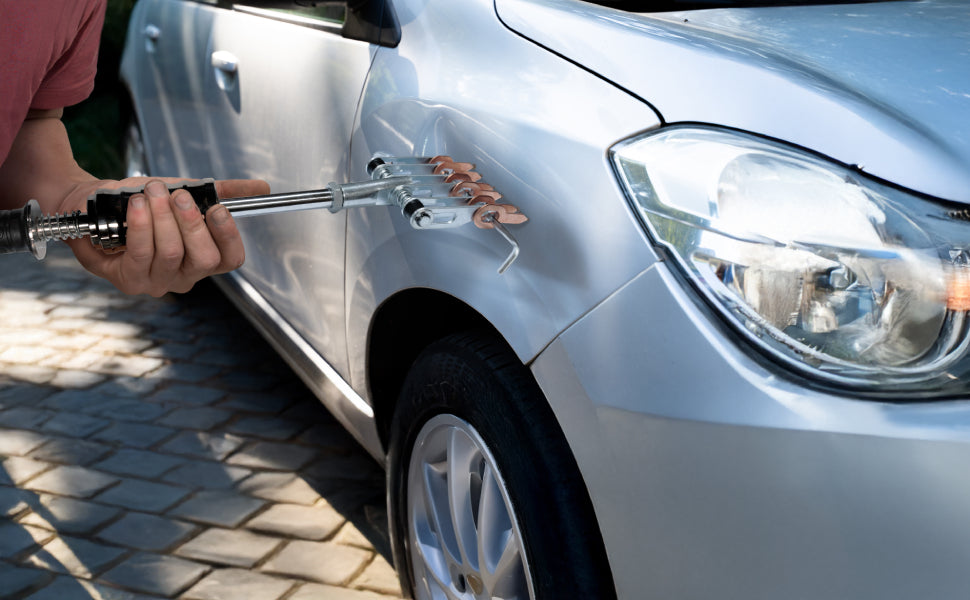 Eine Person repariert eine silberne Autokarosserie mit einem speziellen Werkzeug, das Metallverformungen behebt, auf einem gepflasterten Boden