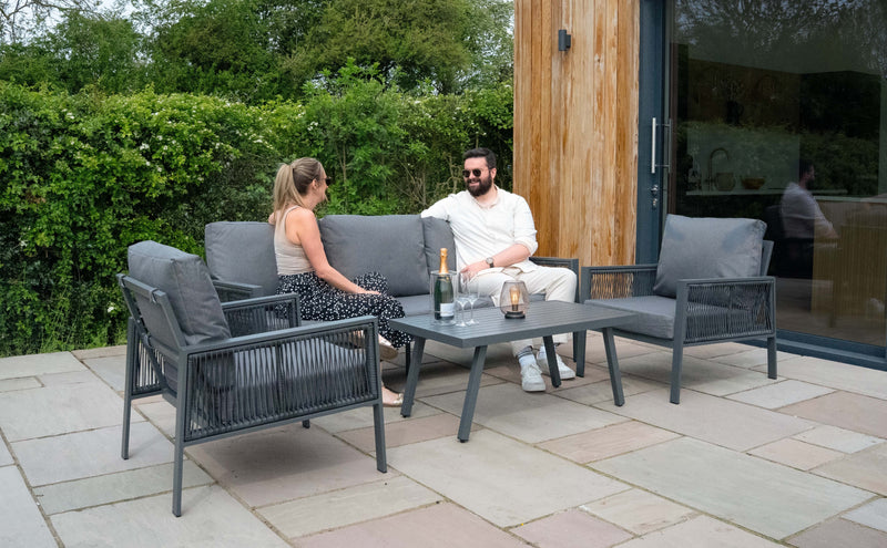 Zwei Personen entspannen auf einer modernen, grauen Gartenlounge mit einem Tisch, umgeben von grünen Pflanzen und einem eleganten, natürlichen Ambiente