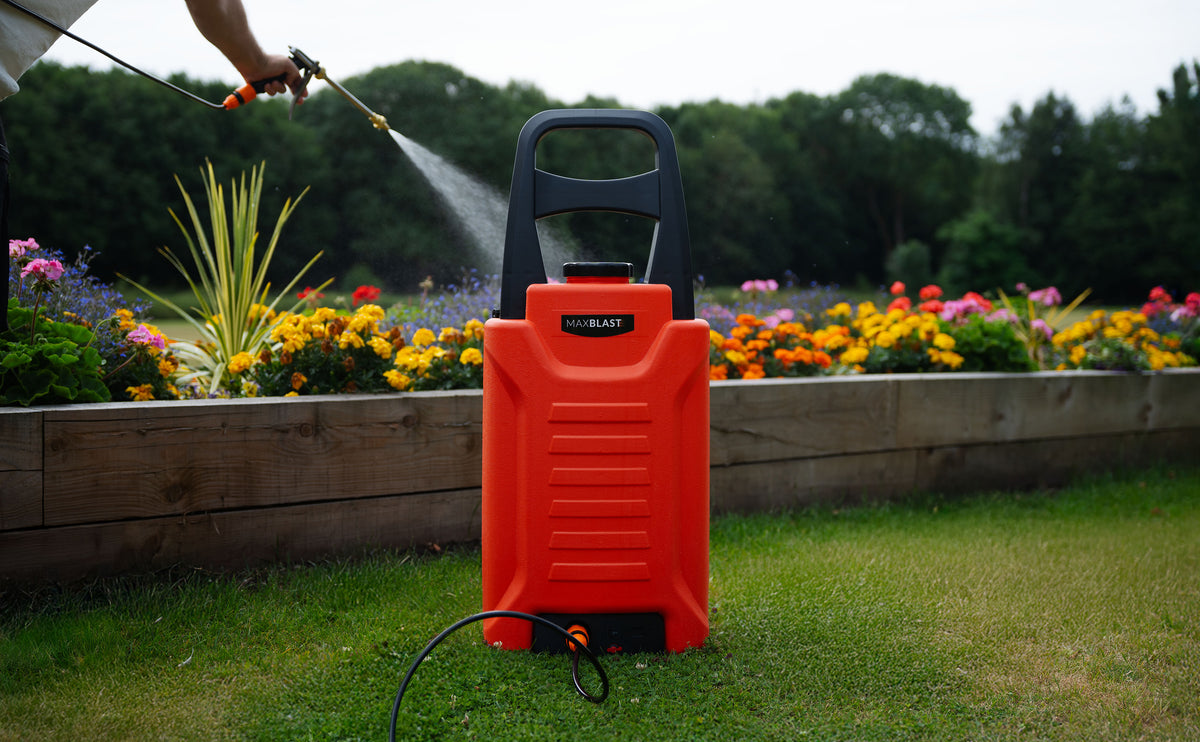 Roter Hochdruckreiniger mit ergonomischem Griff, auf grünem Rasen vor bunten Blumenbeeten, sprüht Wasser in die Luft