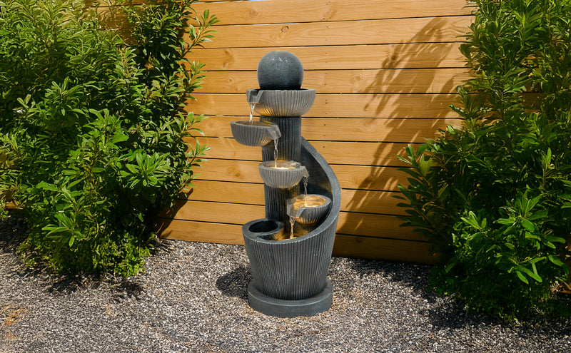 Elegante Gartenbrunnen aus grauem Stein mit mehreren Wasserfällen, umgeben von grünen Pflanzen und einer Holzwand im Hintergrund