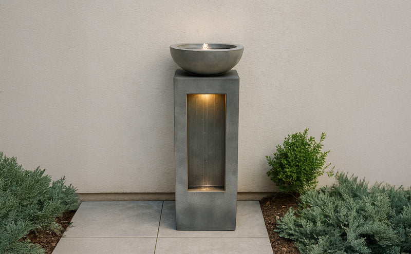 Moderne, graue Betonsäule mit runder Schale oben, die sanft leuchtet; umgeben von grünen Pflanzen und grauen Fliesen, ideal für den Außenbereich
