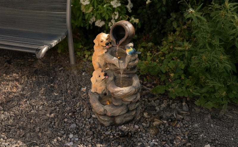 Zwei verspielte Welpen aus Stein sitzen an einem Wasserbrunnen, aus dem Wasser fließt, umgeben von grünen Pflanzen und Kieselsteinen