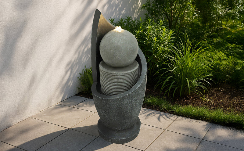 Elegante Gartenbrunnen aus grauem Stein mit einer runden Kugel, die sanft Wasser sprudelt, umgeben von üppigem Grün und modernen Fliesen