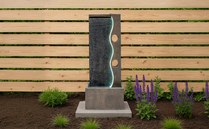 Moderne Gartenbrunnen aus grauem Beton mit geschwungener Wasserlinie, umgeben von lila Blumen und grünen Pflanzen, vor einer Holzlattenwand