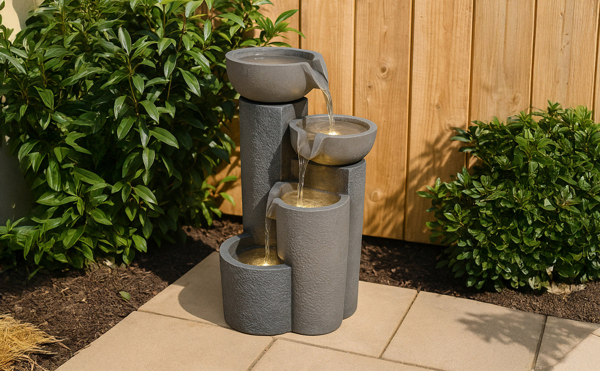 Drei-stufiger Gartenbrunnen aus grauem, strukturiertem Material mit sanft fließendem Wasser, umgeben von grünen Pflanzen und Holzzaun
