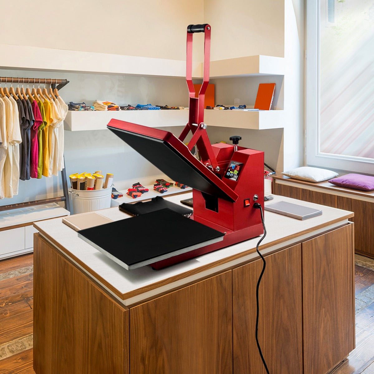 Rote Transferpresse mit schwarzer Heizplatte, auf einem Holztisch in einem modernen Geschäft, umgeben von bunten Textilien und Druckmaterialien