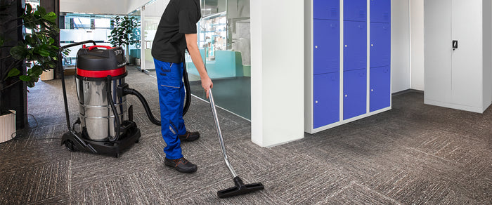 Ein Mann in blauen Arbeitshosen saugt mit einem leistungsstarken Staubsauger den modernen, grauen Teppichboden in einem hellen Büro mit blauen Schränken