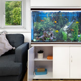 Aquarium Komplettset mit weißem Unterschrank, schwarzem Kies und bunten Fischen, umgeben von grünen Pflanzen und einem gemütlichen Wohnzimmer mit Sofa