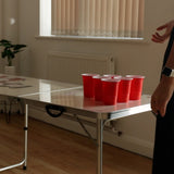 Bier Pong Tisch mit glänzender Oberfläche, sechs roten Plastikbechern auf der Tischkante, Holzfußboden und sanftem Licht im Hintergrund